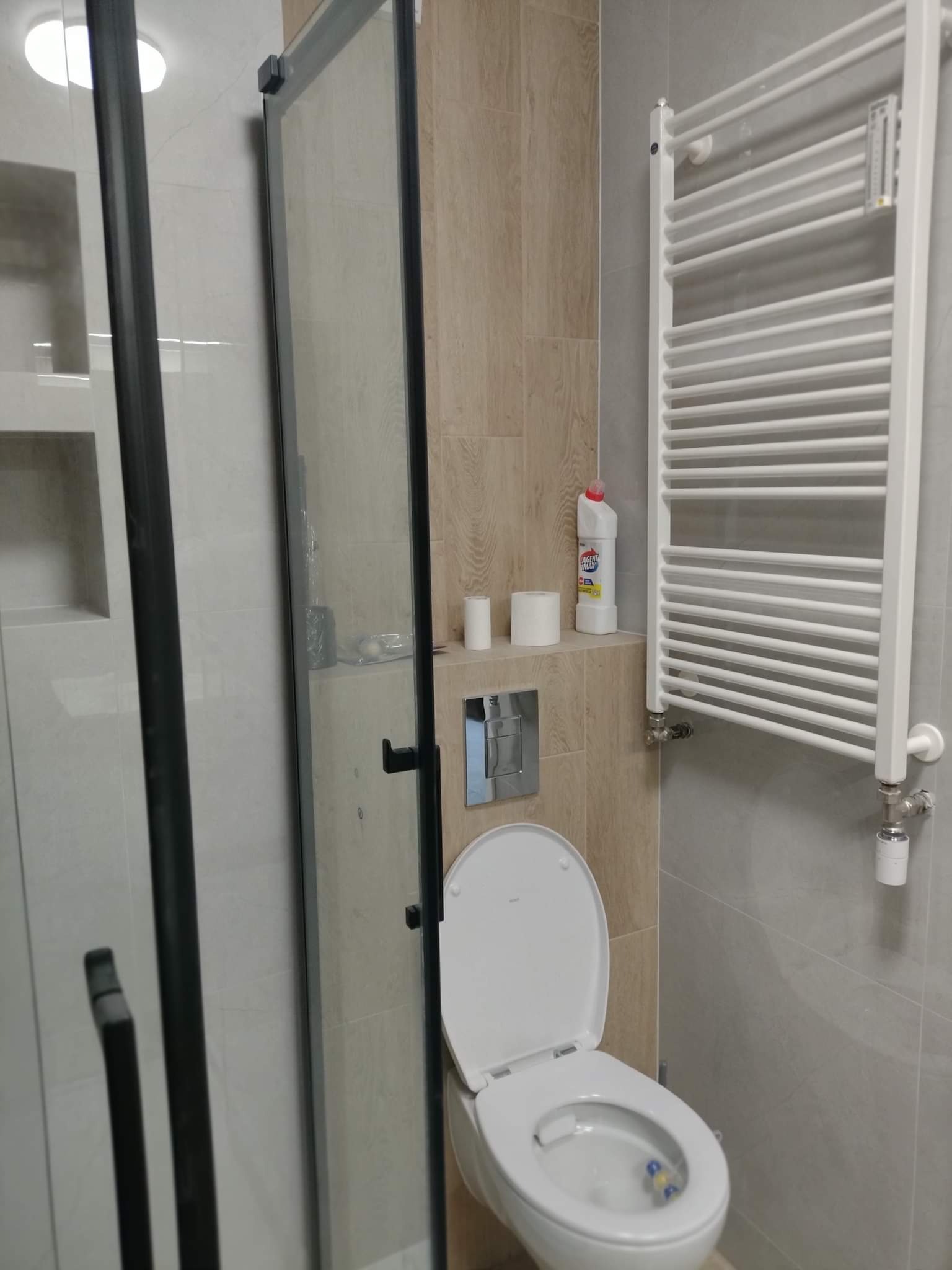 Wykończona łazienka z kabiną prysznicową z czarnymi ramami, toaletą z otwartą klapą, grzejnikiem drabinkowym i półką nad toaletą z kosmetykami.