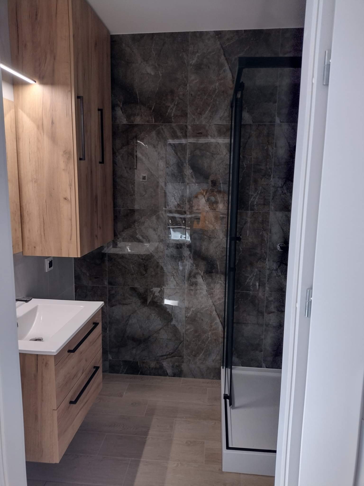 Nowoczesna łazienka z umywalką w szafce z drewnianym fornirem, kabiną prysznicową z czarnymi profilami i marmurowymi płytkami na ścianach.