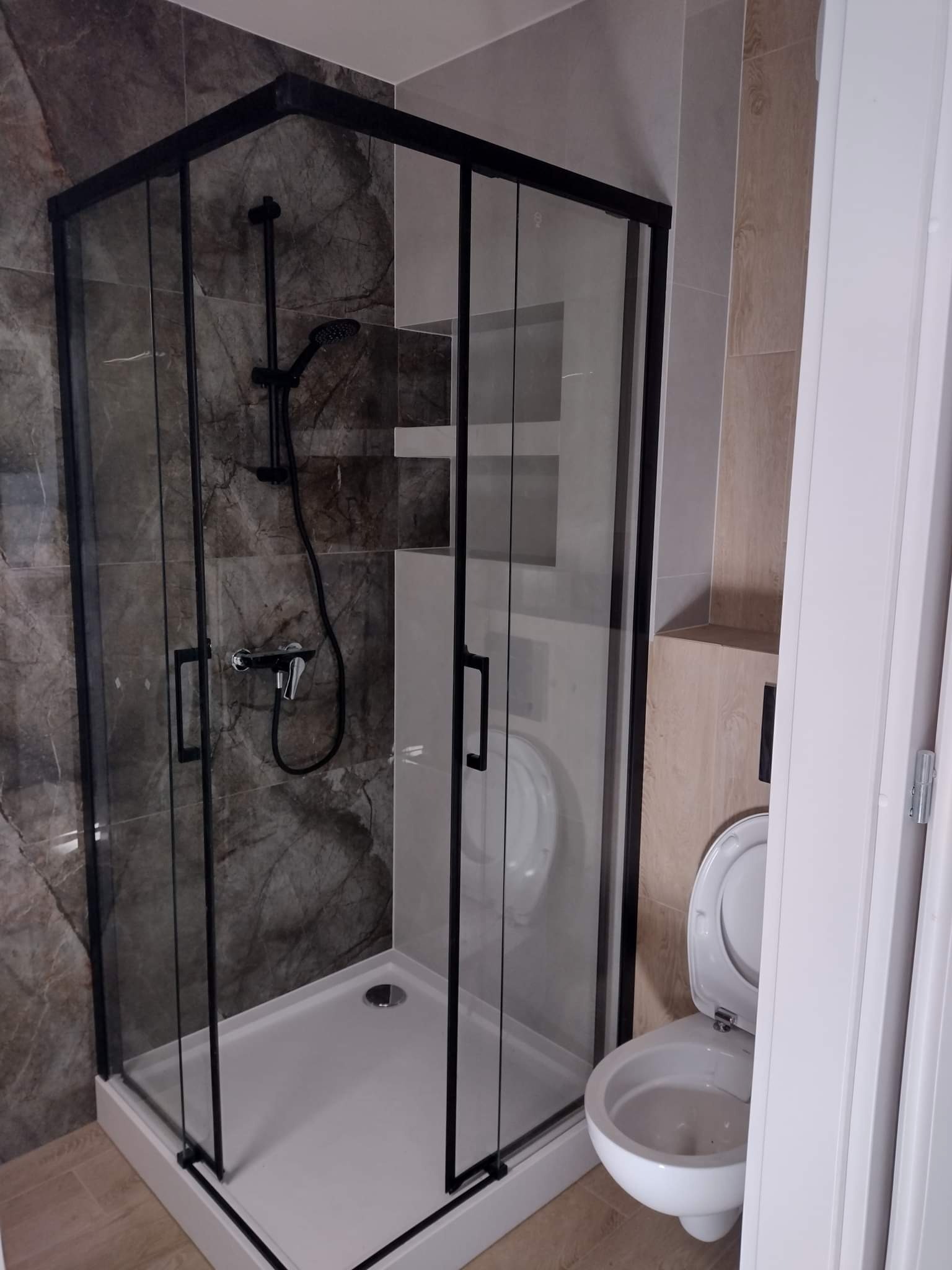 Nowoczesna kabina prysznicowa z czarnymi profilami i deszczownicą na tle marmurowej ściany, obok toaleta z otwartą klapą.