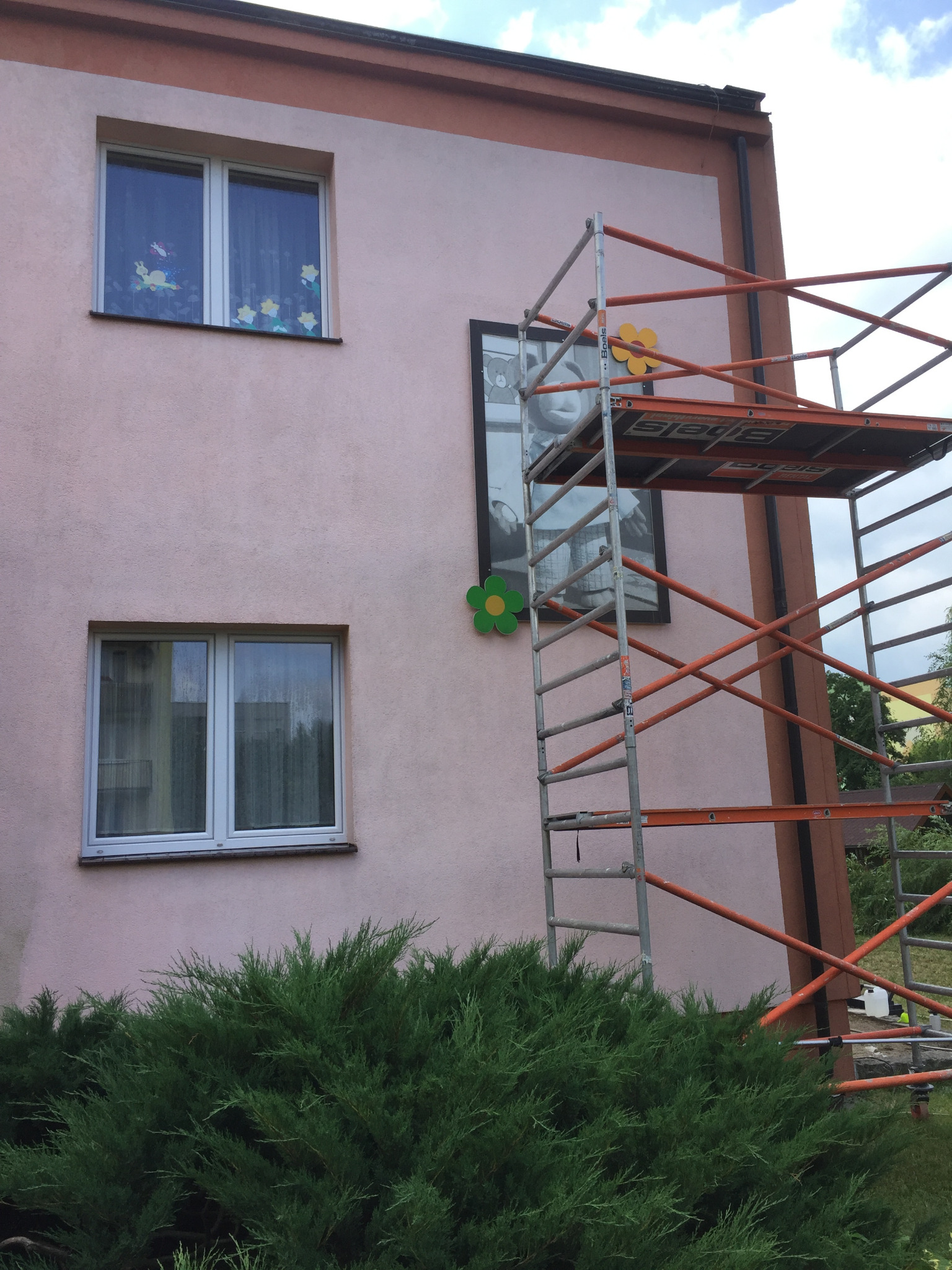 Różowa elewacja budynku przedszkola z rusztowaniem, na którym umieszczono dekoracyjne kwiaty. Okna ozdobione naklejkami z motywami dziecięcymi.