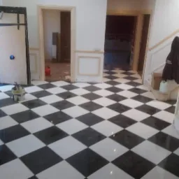 Błyszcząca, czarno-biała podłoga w szachownicę, świeżo po położeniu, z widocznymi niedokończonymi panelami ściennymi i otwartymi drzwiami w tle.