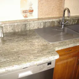 Kuchnia z blatem o zielonkawej barwie i wyraźnym wzorze kamienia, z widocznym zlewozmywakiem, baterią, fragmentem płytek ceramicznych z motywem i częściowo zabudowaną zmywarką Siemens.