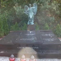 Czarny, wypolerowany pomnik rodzinny z inskrypcjami i rzeźbą anioła na tle zieleni, z trzema zniczami u podstawy.