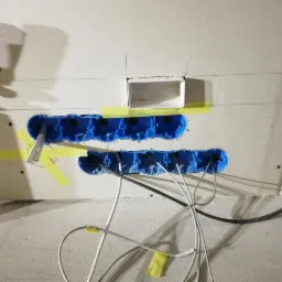 Niebieskie puszki instalacyjne podtynkowe, częściowo oklejone żółtą taśmą, zamontowane w płycie gipsowo-kartonowej z wystającymi przewodami elektrycznymi.