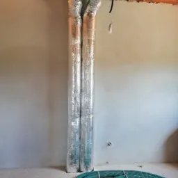 Dwa pionowe przewody wentylacyjne owinięte folią aluminiową, przymocowane do rozdzielacza z zielonymi rurami karbowanymi w surowym stanie budynku.