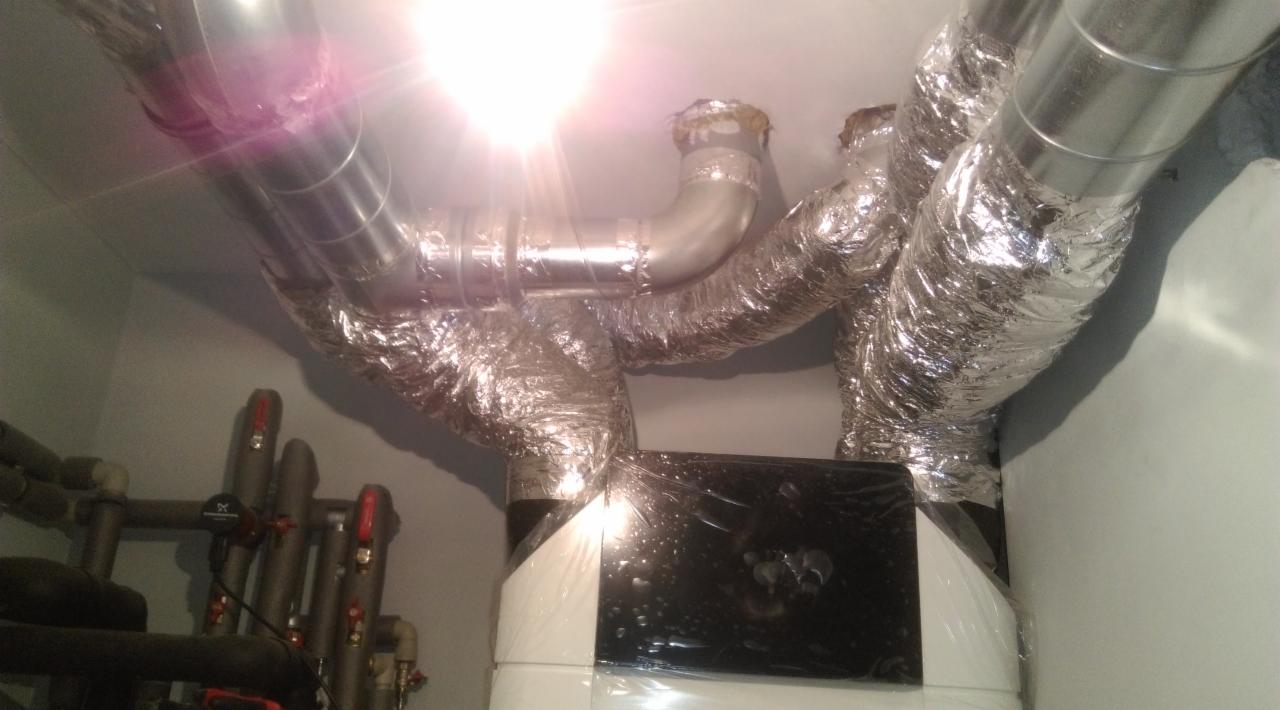 Biała jednostka centralna z czarnym panelem, owinięta folią ochronną, podłączona do srebrnych, izolowanych przewodów wentylacyjnych pod sufitem.