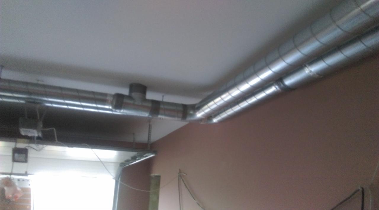 Dwa równoległe, metalowe przewody wentylacyjne przymocowane do sufitu, z widocznymi elementami łączeniowymi i odgałęzieniem w kształcie litery T. W tle widoczna ściana w kolorze brudnego różu...