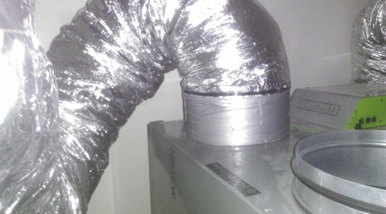 Fragment instalacji: elastyczny, aluminiowy przewód wentylacyjny podłączony do urządzenia, zabezpieczony srebrną taśmą. Widoczny fragment innego przewodu oraz element obudowy urządzenia z zielonym...