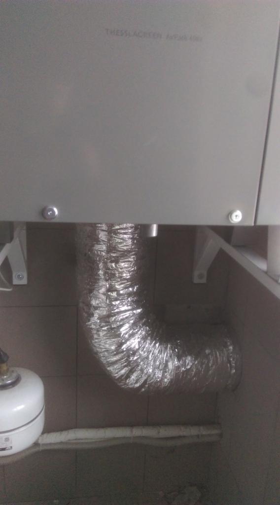 Szary moduł Thessla Green AirPack 4000 z podłączonym elastycznym, aluminiowym przewodem wentylacyjnym w pomieszczeniu z brązowymi płytkami.