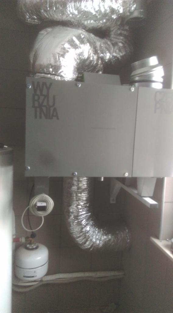 Szara centrala z napisem 'WYRZUTNIA' podłączona do srebrnych, elastycznych przewodów wentylacyjnych, zamontowana w pomieszczeniu z płytkami ceramicznymi.