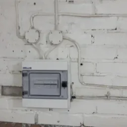 Szczecińska rozdzielnica elektryczna natynkowa, zamontowana na pomalowanej na biało ceglanej ścianie. Widoczne przewody w białych rurkach oraz puszki instalacyjne.