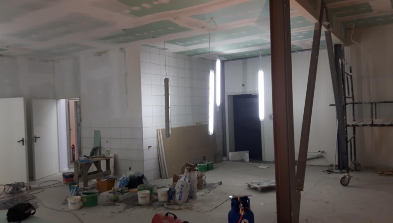 Wnętrze w trakcie remontu z widocznymi lampami wiszącymi, płytami gipsowymi na suficie, narzędziami i materiałami budowlanymi na podłodze, w tle ściany wyłożone białymi płytkami.