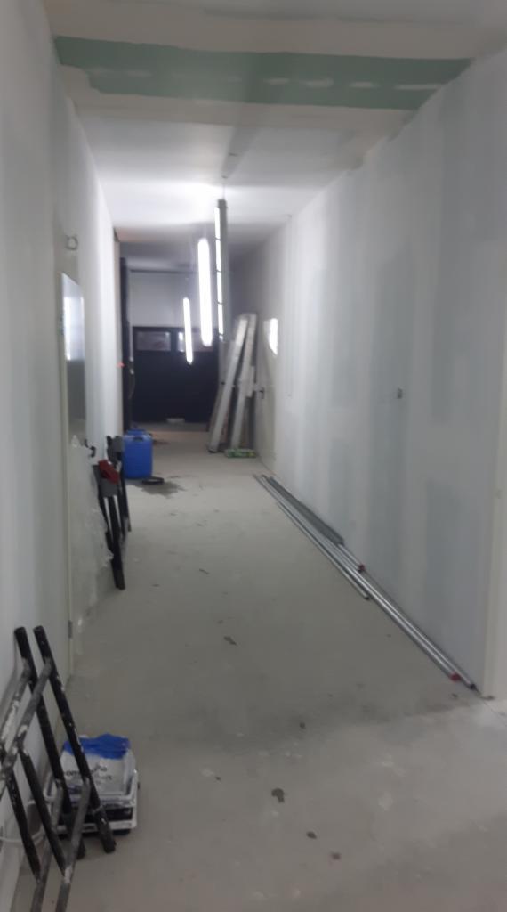 Korytarz w trakcie remontu: surowe ściany przygotowane do malowania, widoczne profile i drabina, oświetlenie tymczasowe.