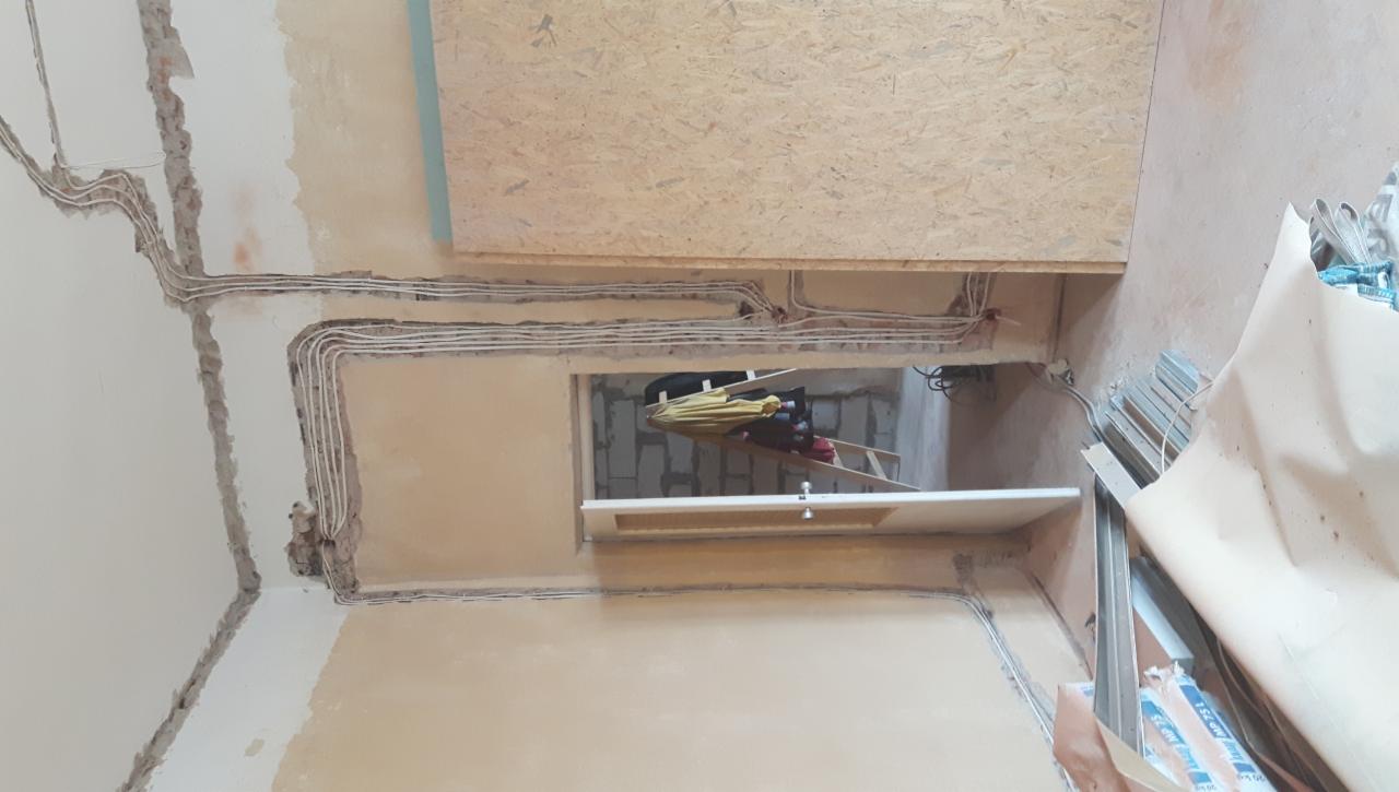 Wnętrze w trakcie remontu z widocznymi przewodami elektrycznymi ułożonymi wzdłuż ścian i sufitu; w tle widoczne rusztowanie i materiały budowlane.