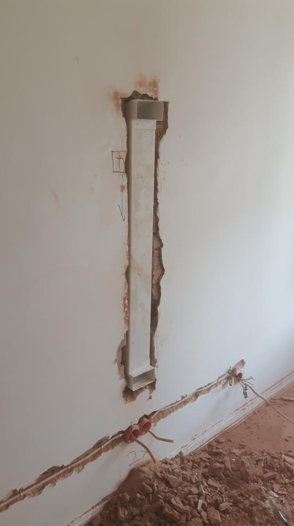 Wykuta bruzda w ścianie z zamontowaną białą rurą osłonową na przewody, widoczne kable w dolnej części ściany i gruz na podłodze.