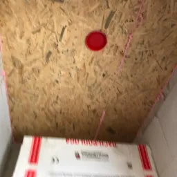 Płyta OSB zamontowana na suficie, uszczelniona różową pianką montażową, widoczna czerwona puszka instalacyjna i fragment opakowania materiału budowlanego.