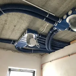 Niebieskie, karbowane rury wentylacyjne podwieszone do surowego betonowego stropu, z widocznymi elementami montażowymi i skrzynkami rozdzielczymi.