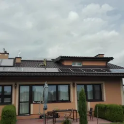 Dom jednorodzinny z brązowym dachem, na którym zamontowane są panele fotowoltaiczne; widoczna antena satelitarna na ścianie budynku i częściowo rozłożony parasol ogrodowy.