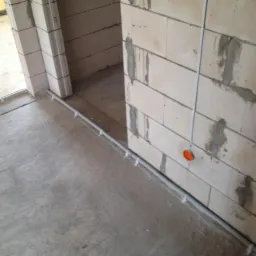 Wnętrze w trakcie budowy z widocznymi przewodami elektrycznymi ułożonymi wzdłuż ściany z bloczków betonowych i podłogi, zabezpieczone plastikowymi uchwytami, z pomarańczową puszką elektryczną...