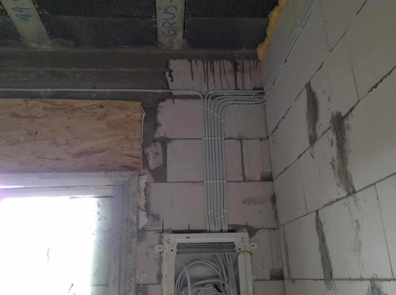 Białe rurki instalacyjne prowadzone po ścianie z bloczków betonowych do otwartej skrzynki rozdzielczej z widocznymi przewodami, surowy stan wykończenia wnętrza.