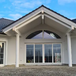 Elewacja domu jednorodzinnego z dużymi przesuwnymi drzwiami tarasowymi pod półkolistym oknem, widoczne okna z białymi ramami i czarnym dachem na tle błękitnego nieba.