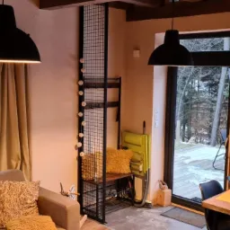 Salon z dużymi, ciemnymi oknami wychodzącymi na ośnieżony las, widoczne czarne ramy okienne i fragment sofy z poduszkami w odcieniach beżu i brązu, wiszące czarne lampy.