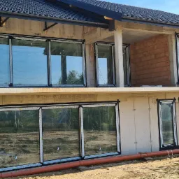 Surowy stan budynku z zamontowanymi oknami z czarnymi ramami zabezpieczonymi folią, widok na wzgórza w tle.