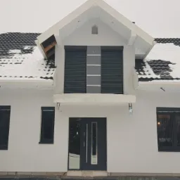 Nowoczesny dom jednorodzinny z szarymi oknami, drzwiami i roletami zewnętrznymi w zimowej scenerii, dach pokryty śniegiem.