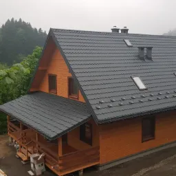Nowoczesny, drewniany dom z szarą, metalową dachówką i oknami dachowymi, usytuowany na tle zalesionych wzgórz w pochmurny dzień.
