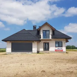 Dom w trakcie budowy z zamontowaną bramą garażową, oknami i drzwiami zewnętrznymi, widoczne białe bloczki i ciemna dachówka, plac budowy z piaskiem i pole w tle.