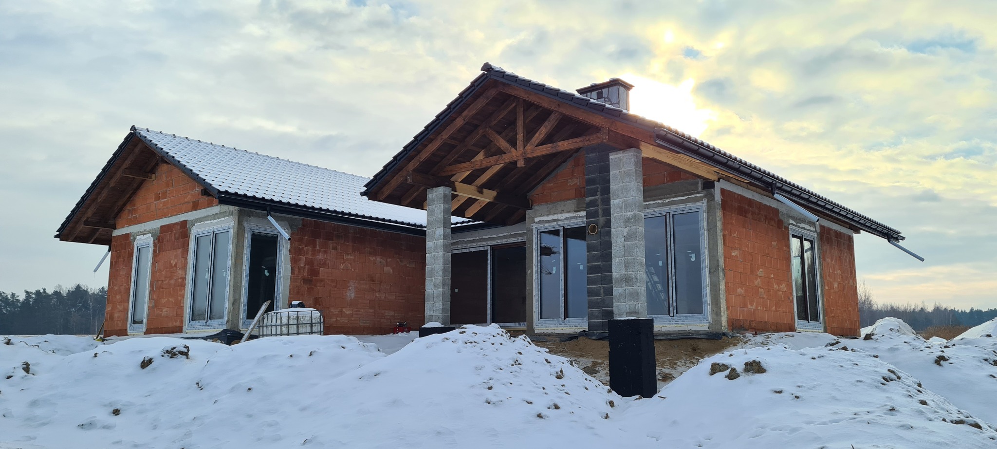 Budowa domu jednorodzinnego w stanie surowym zimą, widoczne zamontowane okna, ceglana elewacja i konstrukcja dachu z drewnianymi elementami.