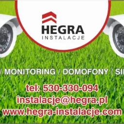 Grafika reklamowa firmy Hegra Instalacje z dwoma białymi kamerami monitoringu na tle zielonej trawy, prezentująca usługi: alarmy, monitoring, domofony i sieci FTTH, wraz z numerem telefonu...