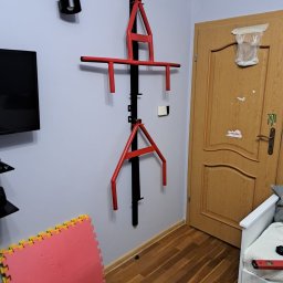 Metalowe Fantazje - Czerwono-czarny, regulowany drążek do podciągania zamontowany na szarej ścianie w pokoju, widoczne puzzle piankowe na podłodze i drzwi w tle.