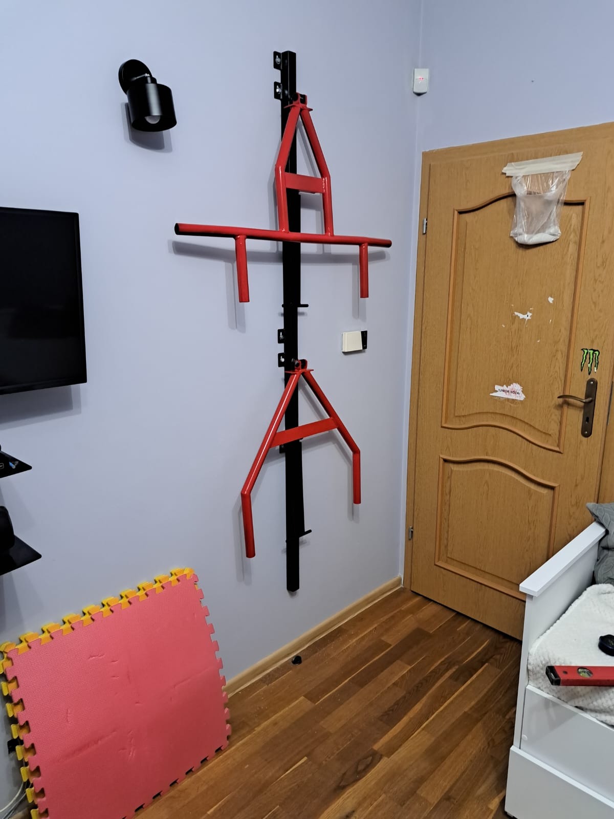 Czerwono-czarny, regulowany drążek do podciągania zamontowany na szarej ścianie w pokoju, widoczne puzzle piankowe na podłodze i drzwi w tle.