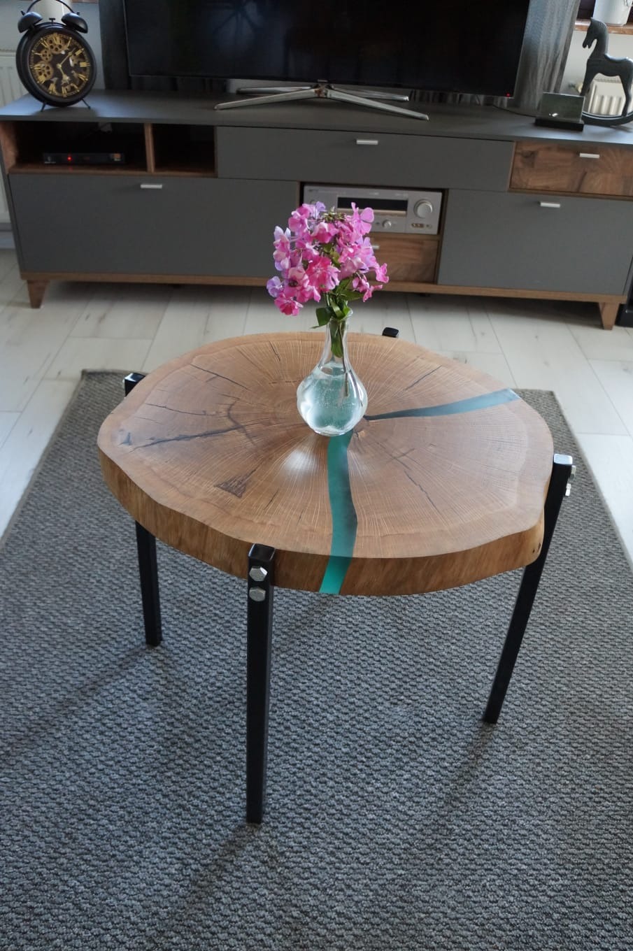 Okrągły stolik kawowy z blatem z plastra dębu wypełnionego turkusową żywicą epoksydową i wazonem z różowymi kwiatami, na tle szarej szafki RTV i dywanu.