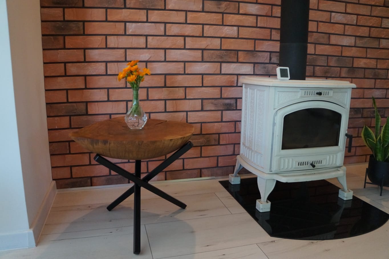 Stylowy piec wolnostojący w kolorze złamanej bieli z czarnym kominem, ustawiony na czarnej, błyszczącej płycie podłogowej w Gorzowie Wielkopolskim, obok stolik z blatem z litego drewna i bukietem...