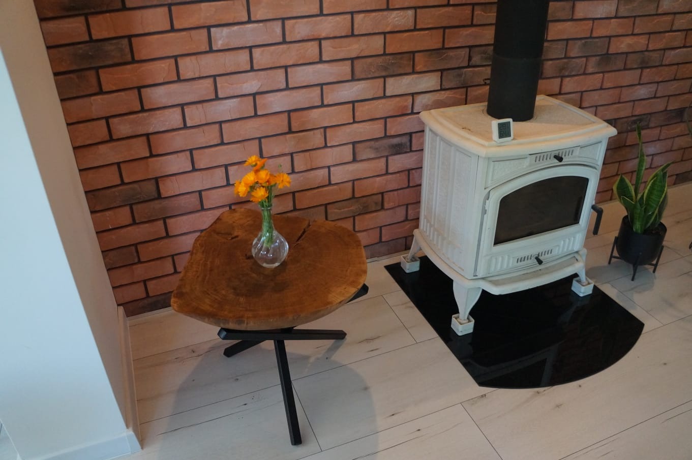 Stylowy salon z piecem wolnostojącym w kolorze białym, na podłodze jasne panele, a na ścianie dekoracyjna cegła. Obok pieca stolik kawowy z blatem z plastra drewna i bukietem pomarańczowych kwiatów...