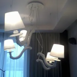 Biały, ozdobny żyrandol z abażurami w trakcie montażu, widoczne kable i mocowanie do sufitu.