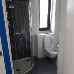 Nowa łazienka w trakcie wykańczania: kabina prysznicowa z chromowanymi detalami, podwieszana toaleta z deską w folii ochronnej, czarne heksagonalne płytki podłogowe, białe ściany i czarna rama...