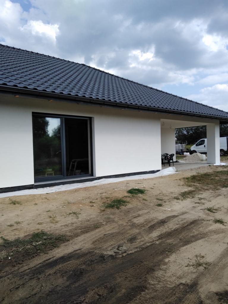 Parterowy dom z ciemnym dachem i białymi ścianami po remoncie. Widoczny fragment elewacji, okno i zadaszone wejście. Tło: pochmurne niebo.