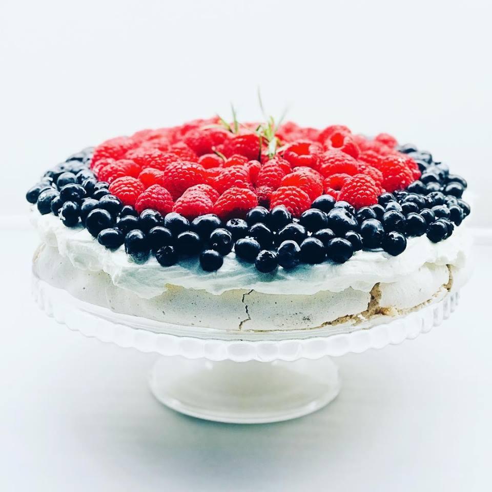 Tort bezowy udekorowany świeżymi malinami i borówkami, na szklanej paterze z nóżką, z gałązką rozmarynu jako ozdobą.