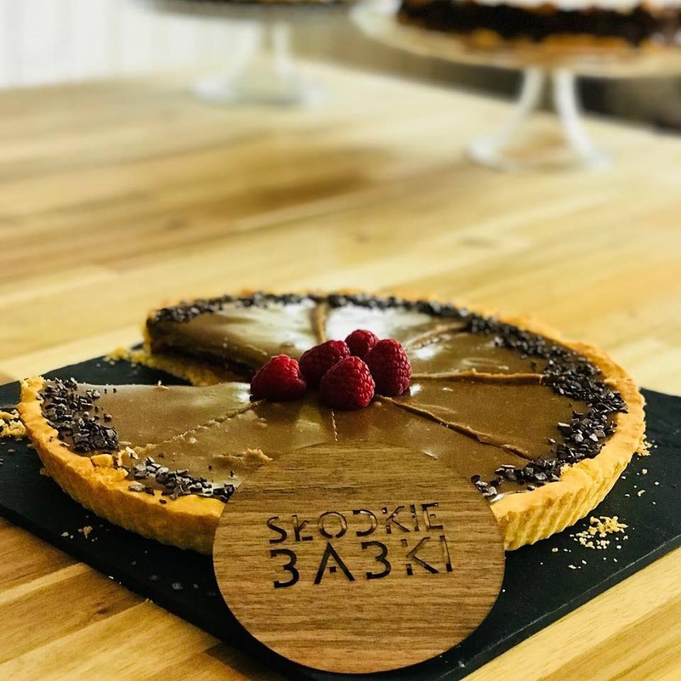 Tartaletka z czekoladowym kremem, malinami i posypką czekoladową, prezentowana na czarnym talerzu z drewnianym logo 'Słodkie 3A3KI'. W tle widoczne inne ciasta na paterach.