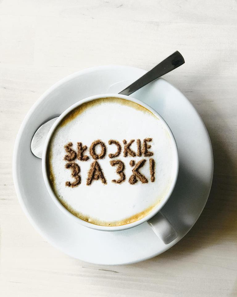Cappuccino z napisem 'Słodkie 3A3KI!' wykonanym z kakao, podane na białym spodku z łyżeczką.