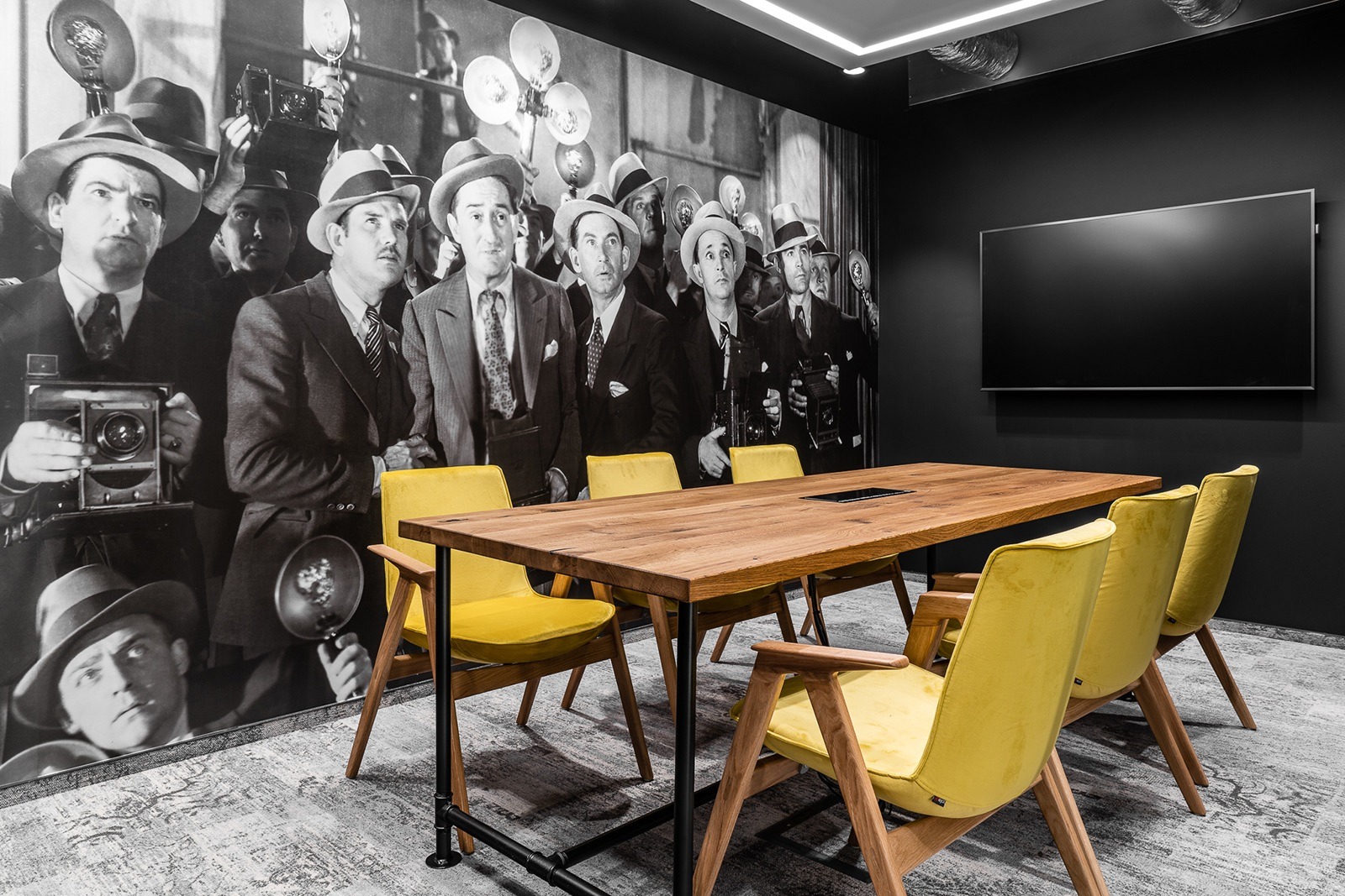 Nowoczesna sala konferencyjna z drewnianym stołem i żółtymi krzesłami, na ścianie fototapeta z motywem retro fotografów i duży ekran. Stonowana kolorystyka.
