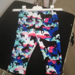 Dziecięce legginsy z motywem czarnych jednorożców na białym tle z plamami koloru niebieskiego i różowego, wiszące na białym wieszaku nad czarnym obiciem krzesła.