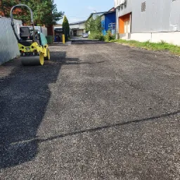 Żółty walec drogowy AMMANN na świeżo utwardzonej, ciemnej nawierzchni z kruszywa, w tle budynki przemysłowe i zieleń.