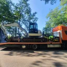 Pomarańczowa laweta z minikoparką i walcem drogowym na platformie; pracownik w ż&oacute;łtej kamizelce zabezpiecza ładunek pasami; drzewa w tle.