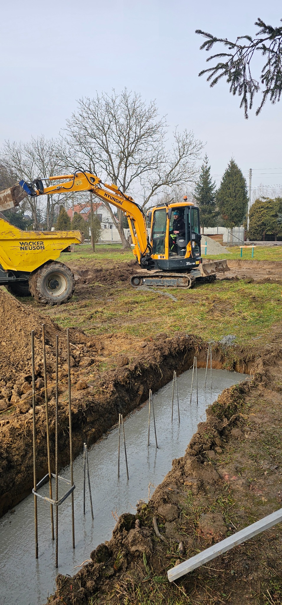 Żółta koparka Hyundai i wywrotka Wacker Neuson obok wykopu z zalanym betonem i wystającymi prętami zbrojeniowymi na placu budowy.