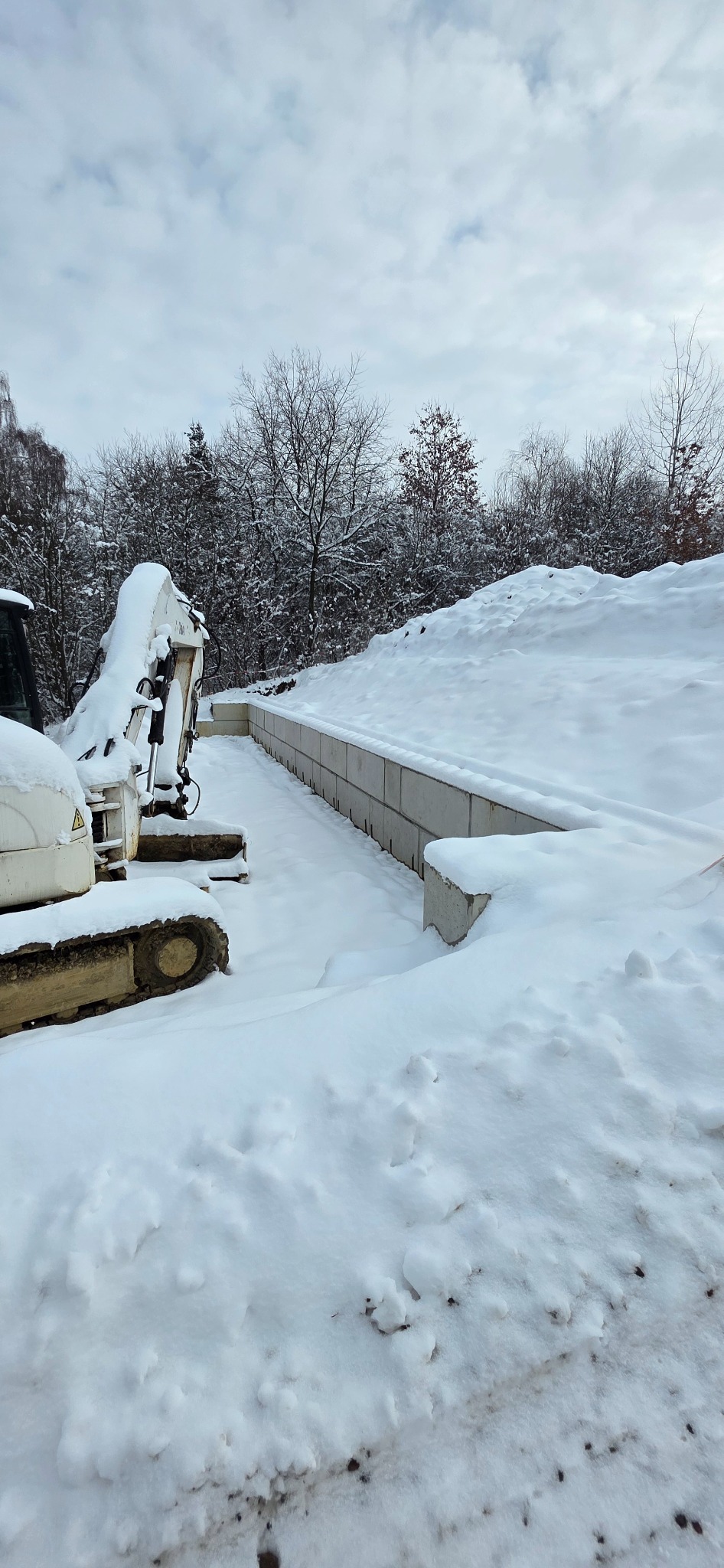 Śnieżny krajobraz z wykopem zabezpieczonym betonowymi blokami i częściowo widoczną białą koparką. W tle drzewa pokryte śniegiem i pochmurne niebo.