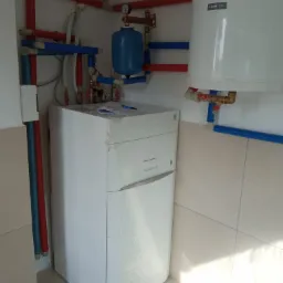 Biały piec grzewczy De Dietrich w folii ochronnej, podłączony do instalacji z rur miedzianych i plastikowych (czerwone i niebieskie), z niebieskim zbiornikiem wyrównawczym i białym zasobnikiem...